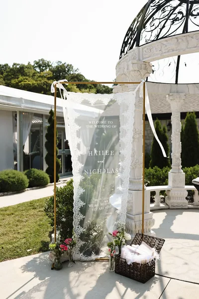 An acrylic welcome sign draped in lace, standing beside the ceremony gazebo.