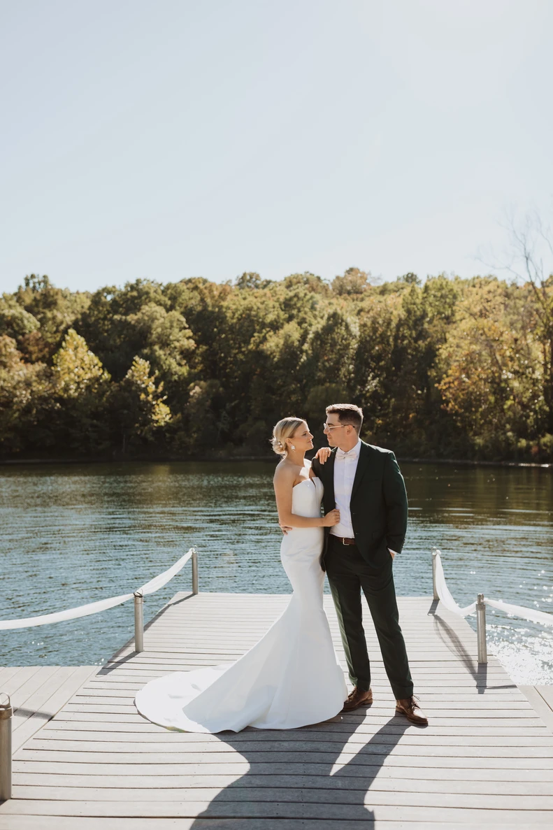 A bride and groom stand together at the end of a lake dock in the afternoon sun, calm water and a tree line behind them.