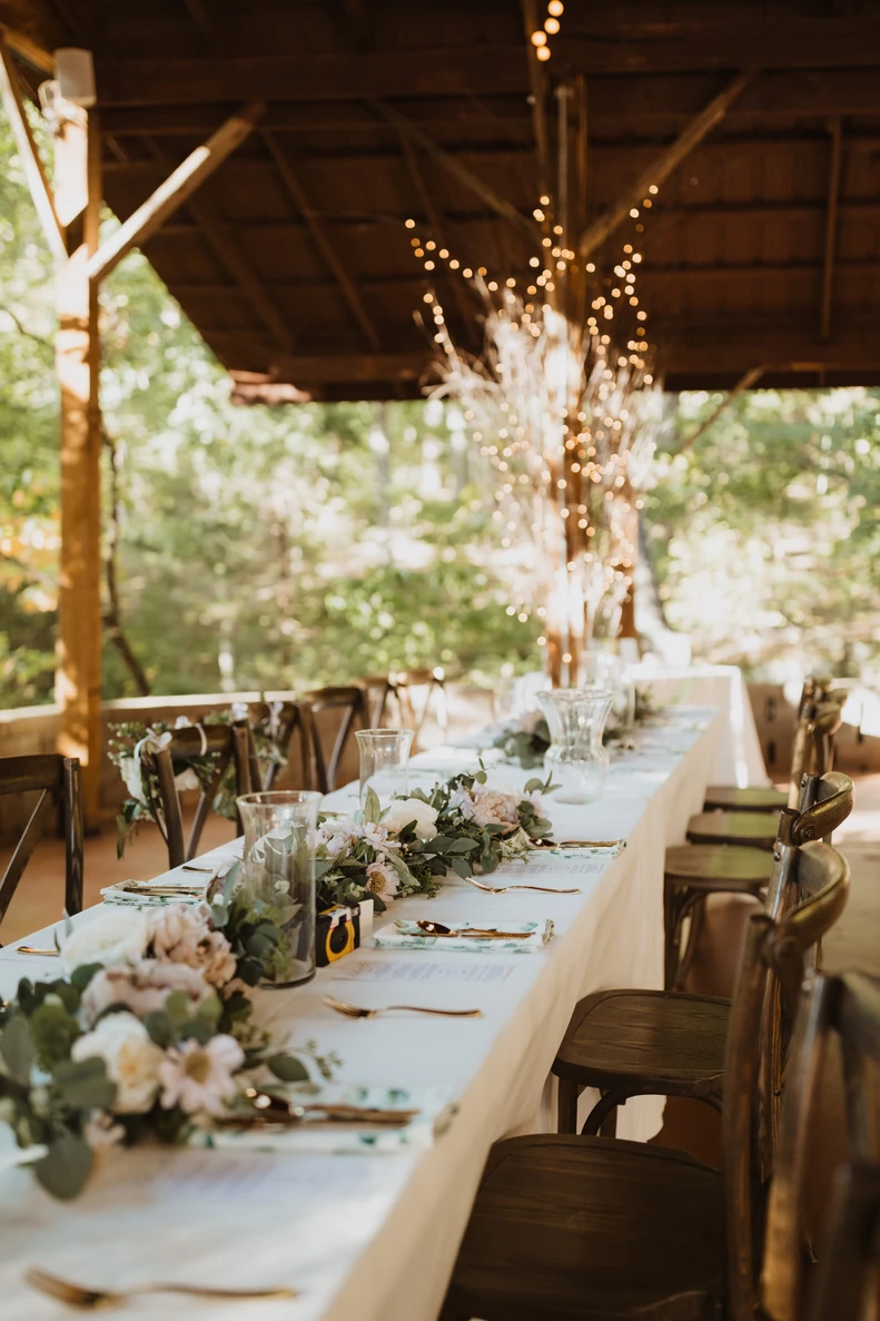A long head table set under string lights, dressed with white florals and greenery, ready for the reception.