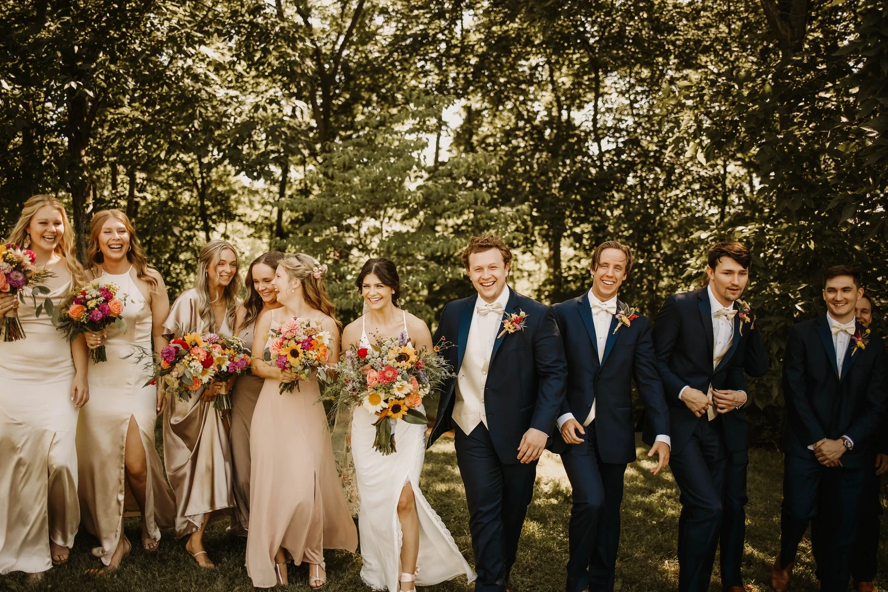 A joyful wedding party walks together through a sunlit tree line, bouquets of sunflowers and wildflowers in hand.