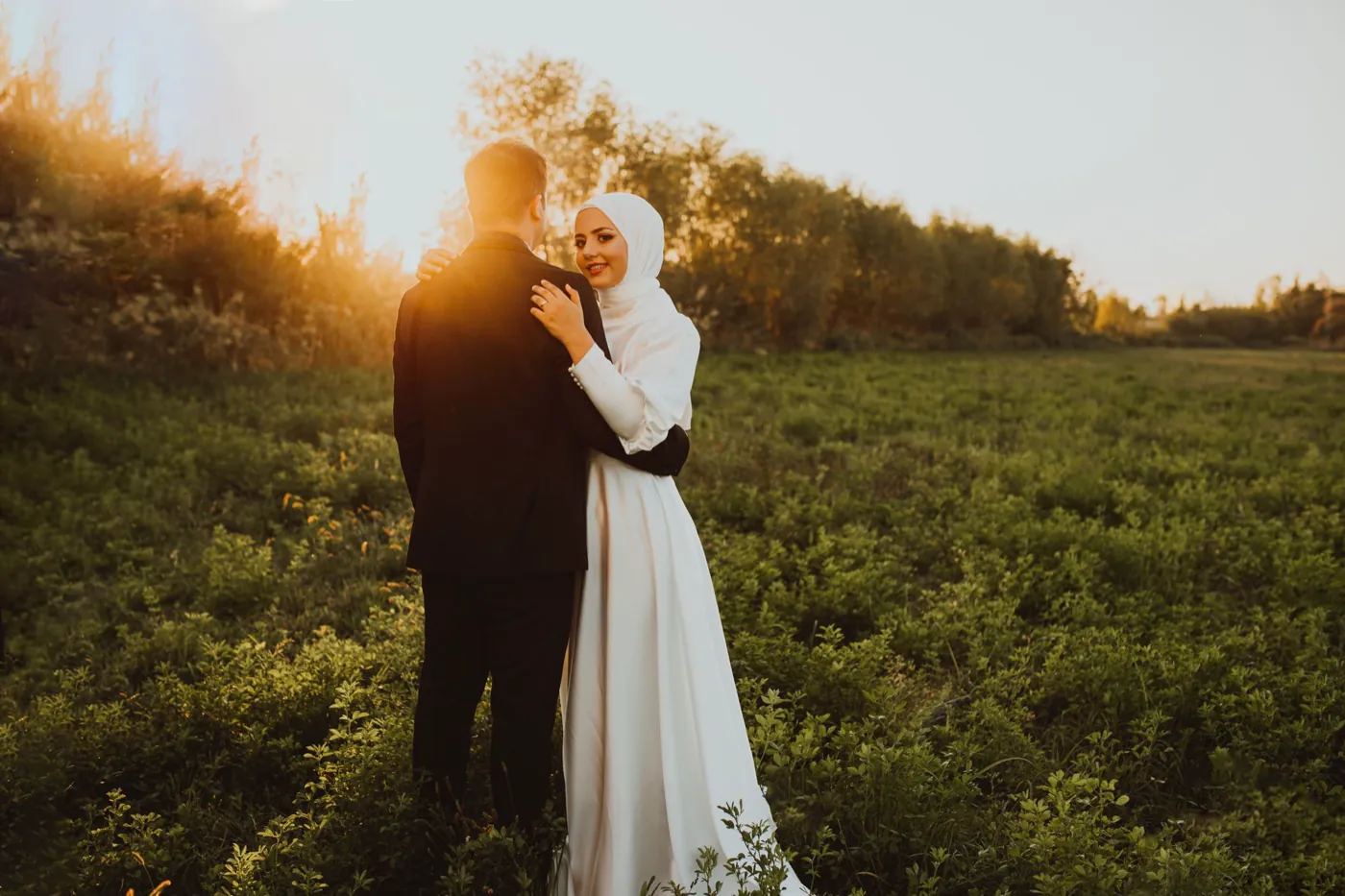 A couple embraces in a green field at golden hour, warm sun flaring behind them.