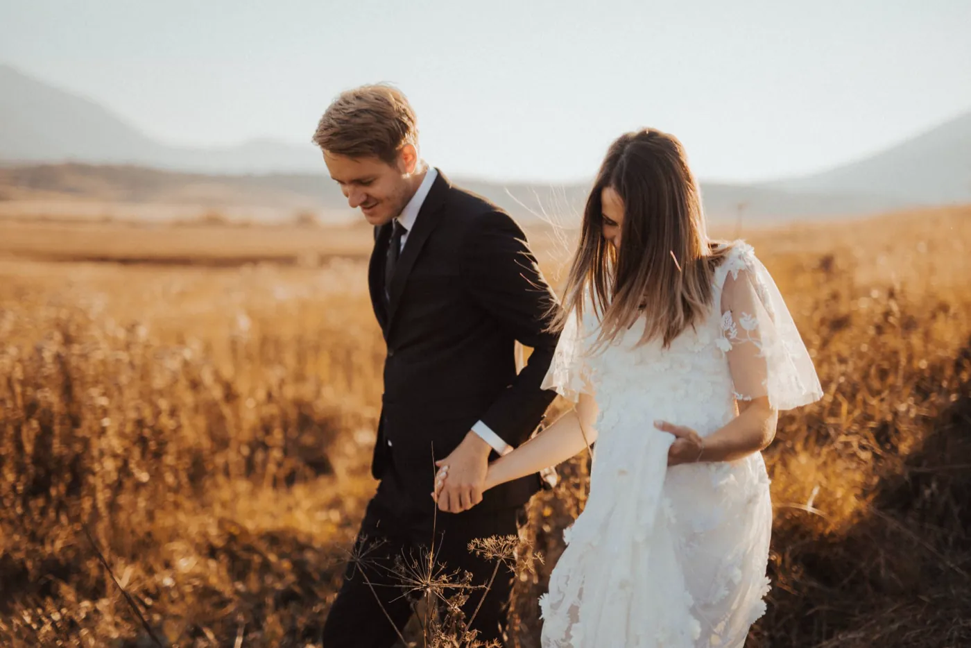 A bride and groom walk hand in hand through a golden wheat field at sunset.
